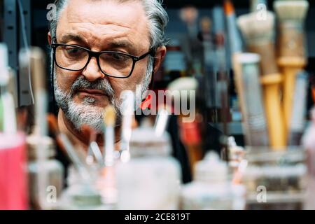 artista maturo concentrato in occhiali lavorando dietro la collezione di pennelli di diverse dimensioni e bottiglie con vernice in studio creativo Foto Stock