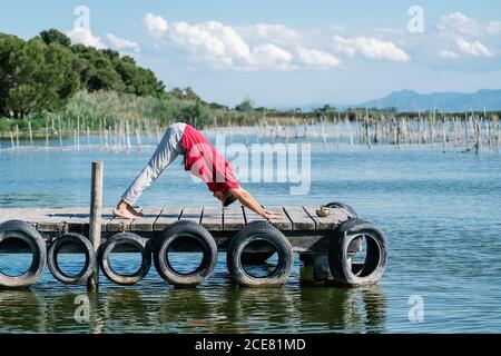 Corpo pieno di un ragazzo tranquillo barefooted in esecuzione di abbigliamento casual yoga posa sul molo di legname in giornata di sole Foto Stock