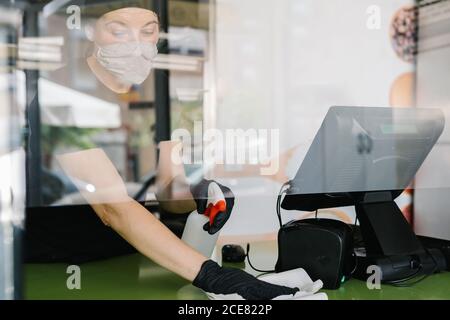Attraverso il raccolto di vetro concentrato femmina cassiere indossando respiratore e lattice guanti che strofinano il contatore con il panno mentre si lavora in un bar a. prevenire la diffusione del coronavirus Foto Stock