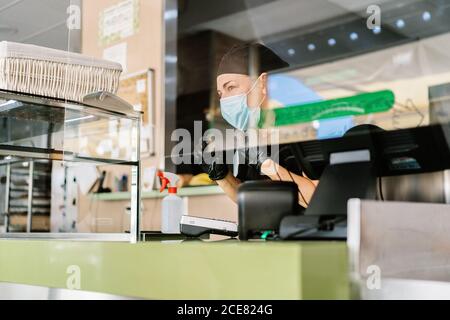 Il personale del caffè è molto positivo in uniformi e respiratori che lavorano al bar contatore con registratore di cassa durante la pandemia di coronavirus Foto Stock