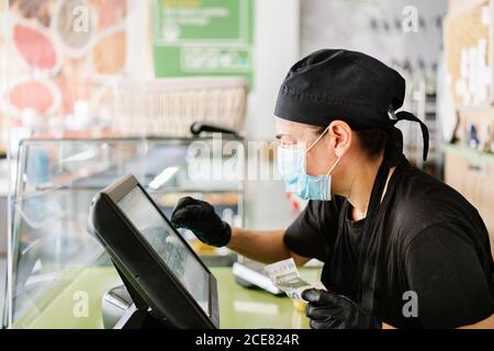 Vista laterale cassiere femminile grave con guanti e maschiatura sul registratore di cassa touchscreen durante il servizio in una caffetteria contemporanea Foto Stock