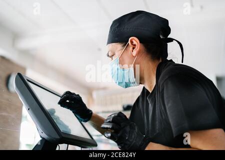 Vista laterale cassiere femminile grave con guanti e maschiatura sul registratore di cassa touchscreen durante il servizio in una caffetteria contemporanea Foto Stock