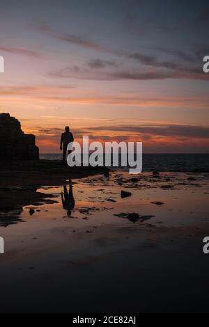 anonimo viaggiatore maschile in piedi vicino al monte e mare infinito sotto cielo spettacolare al tramonto che si riflette in acqua Foto Stock