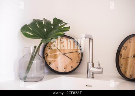 Set di orologio rotondo e foglia verde di monstera in vetro vaso disposto su lavandino vicino a parete bianca in cucina leggera Foto Stock