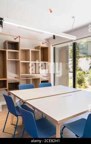 Interni in stile moderno con tavoli quadrati e sgabelli blu su pavimento e ripiani in legno su parete con grande finestra e lampade sul soffitto Foto Stock