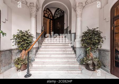 Grande scala con ringhiera in legno e colonne classiche con ornamento fregi vicino porta ad arco aperto e piante verdi su ceramica pavimento Foto Stock