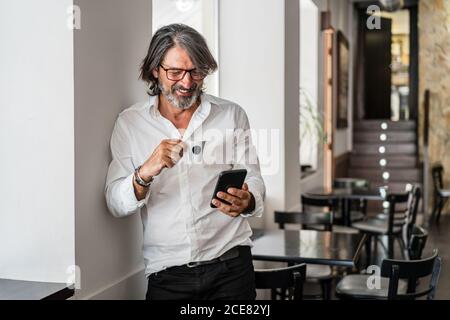 Positivo etnico invecchiato grigio peloso uomo browser smartphone e. avere un caffè mentre si appoggia al muro in un caffè vuoto giorno Foto Stock