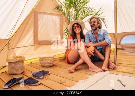 Coppia felice rilassata in abiti casual seduti con gambe incrociate su pavimento in legno in tenda di lusso campeggio durante la spesa vacanze insieme Foto Stock