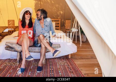 Coppia felice rilassata in abiti casual seduti con gambe incrociate su pavimento in legno in tenda di lusso campeggio durante la spesa vacanze insieme Foto Stock