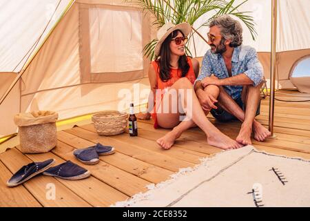 Coppia felice rilassata in abiti casual seduti con gambe incrociate su pavimento in legno in tenda di lusso campeggio durante la spesa vacanze insieme Foto Stock