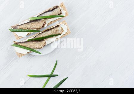 Fresco estivo salutare antipasto di pane intero grattugiato con conserve di spratti, cipolla verde, crema di formaggio su tavola di legno bianco, vista dall'alto, spazio copia Foto Stock