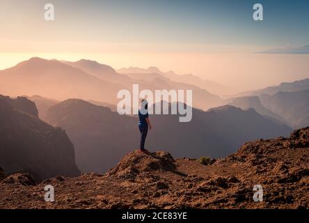 Vista posteriore di anonimo turista femminile in piedi su roccia e. Goditi il magnifico paesaggio di silhouette di montagna durante il tramonto a Gran Canaria Foto Stock