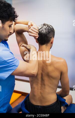 Vista posteriore dell'ortopedico in uniforme controllo del giunto a spalla di calzare un uomo seduto sul lettino di esame con braccio raggiunto in ospedale e guardando via Foto Stock