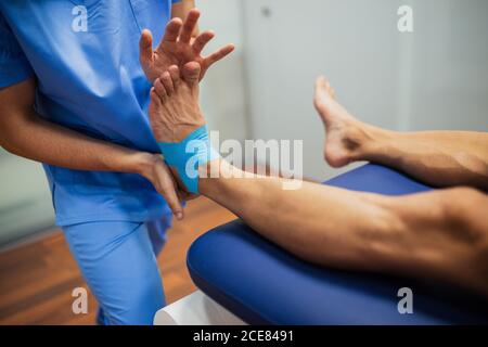 Vista dall'alto del chiropratico irriconoscibile del raccolto con attacco uniforme blu cinesio nastro ai piedi di uomo senza volto che giace all'esame lettino in clinica Foto Stock