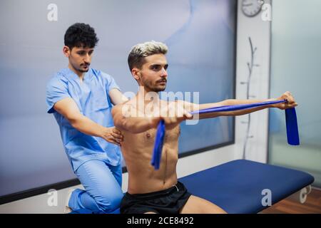 Medico di medicina di sport che controlla la parte posteriore di uomo sportivo con capelli tinti che allungano il nastro elastico durante il periodo di riabilitazione e l'aspetto via Foto Stock