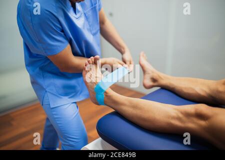 Vista dall'alto del chiropratico irriconoscibile del raccolto con attacco uniforme blu cinesio nastro ai piedi di uomo senza volto che giace all'esame lettino in clinica Foto Stock