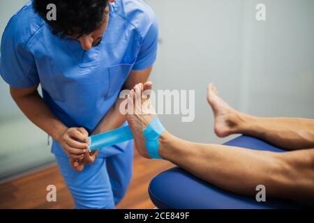 Vista dall'alto del chiropratico irriconoscibile del raccolto con attacco uniforme blu cinesio nastro ai piedi di uomo senza volto che giace all'esame lettino in clinica Foto Stock