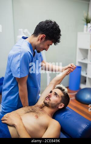 Osteopata nel braccio di esame uniforme blu di un paziente maschile sottile con i capelli tinti giacenti sul tavolo di esame in clinica Foto Stock