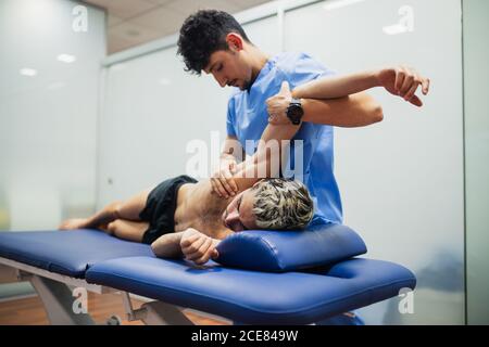 Osteopata con maschera in blu uniforme braccio di esame di sottile paziente di sesso maschile con maschera e capelli tinti giacenti all'esame tavolo in clinica Foto Stock