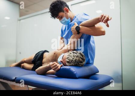 Osteopata con maschera in blu uniforme braccio di esame di sottile paziente di sesso maschile con maschera e capelli tinti giacenti all'esame tavolo in clinica Foto Stock