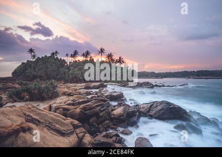 Incredibile scenario di costa rocciosa e palme verdi vicino mare sotto il pittoresco cielo del tramonto Foto Stock