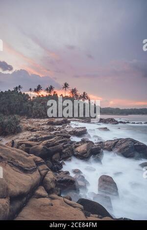 Incredibile scenario di costa rocciosa e palme verdi vicino mare sotto il pittoresco cielo del tramonto Foto Stock