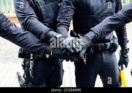 poliziotti anonimi in attrezzi protettivi e maschere mediche armate con pistole e baton che mettono insieme le mani durante la preparazione funzionamento Foto Stock