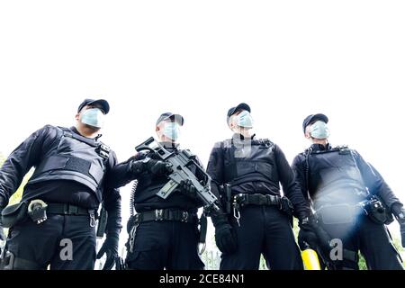 Squadra a corpo pieno di poliziotti spagnoli in attrezzi protettivi con pistole che indossano maschere mediche durante la pattugliatura della strada Foto Stock