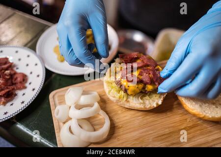 Angolo alto di chef anonimo in guanti medici aggiungendo tritato carne su panino hamburger con cotoletta e verdure Foto Stock