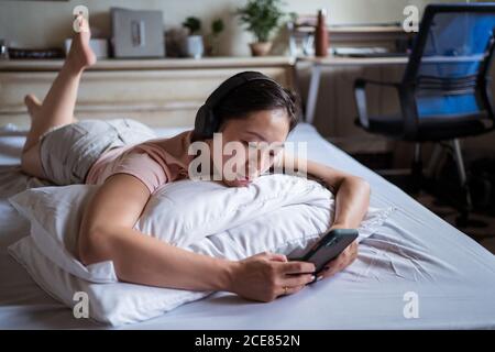 Unhappy Ethnic Woman sdraiato sul letto e utilizzando lo smartphone in una stanza accogliente a casa Foto Stock