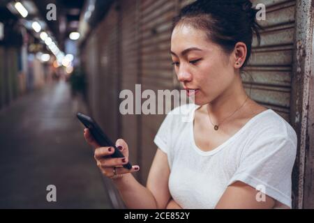 Giovane donna asiatica a corpo pieno in abiti casual con mobile telefono in mano a piedi su un percorso pedonale stretto sotterraneo con negozi chiusi in città Foto Stock