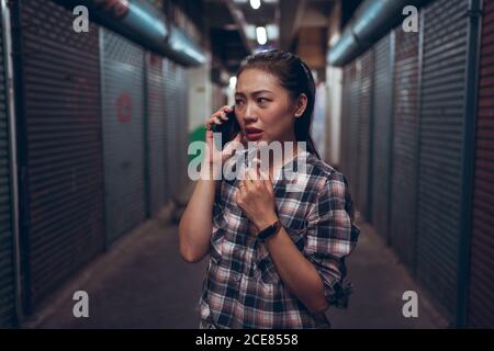 Infelice paura giovane donna asiatica in abbigliamento casual che hanno chiamata sul telefono cellulare mentre si è in piedi in un corridoio sotterraneo scuro grunge Foto Stock