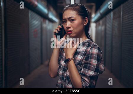 Infelice paura giovane donna asiatica in abbigliamento casual che hanno chiamata sul telefono cellulare mentre si è in piedi in un corridoio sotterraneo scuro grunge Foto Stock