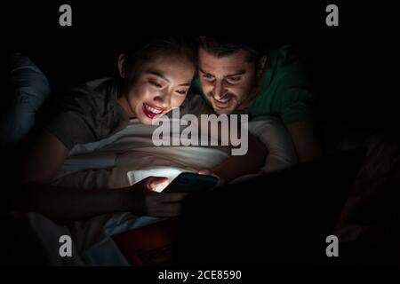 Giovane donna asiatica allegra utilizzando i social media sul cellulare, mentre sdraiato con felice ragazzo a letto di notte in appartamento Foto Stock