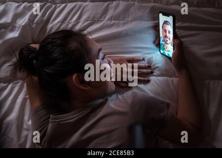 Dall'alto di allegra giovane donna asiatica che parla con bearded amato sul cellulare mentre si effettua la videochiamata e sdraiato in letto di notte a casa Foto Stock
