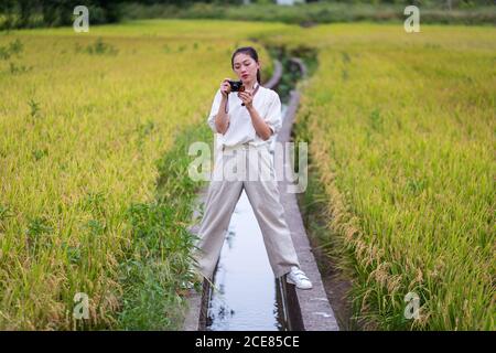 Calma etnica femmina in piedi in campo di riso e scattare foto su una fotocamera vintage Foto Stock