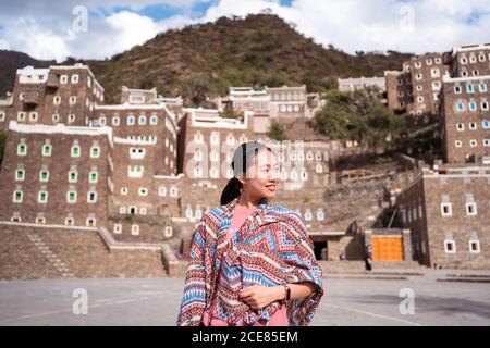 Donna turistica in piedi sullo sfondo di edifici in pietra con colorato finestre mentre si viaggia intorno Rijal Almaa villaggio in Arabia Saudita Foto Stock