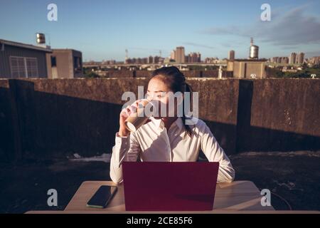 Giovane donna asiatica pensiva in abbigliamento formale seduto a tavola con computer portatile e smartphone e bere caffè da asporto mentre lo si ha pausa durante i lavori in remoto sulla terrazza panoramica Foto Stock