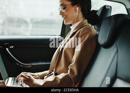 Vista laterale di una donna d'affari sorridente che lavora su un computer portatile mentre viaggia in taxi. Donna che usa il laptop sul sedile posteriore dell'auto. Foto Stock
