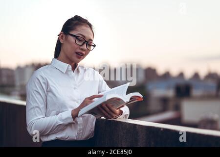 Vista laterale della giovane donna d'affari asiatica con occhiali in abiti formali che si ergono sul tetto dell'edificio cittadino e leggendo un libro mentre si rilassa dopo il lavoro Foto Stock