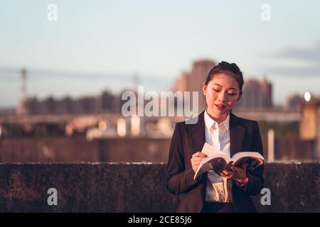 Vista frontale della giovane donna d'affari asiatica in abiti formali che si ergono sul tetto dell'edificio cittadino e leggono un libro mentre si rilassano dopo il lavoro Foto Stock