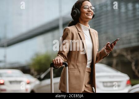 Donna in uomo d'affari che ordina un taxi online con i suoi bagagli mentre si trova in piedi su una strada. Donna sorridente viaggiatore con cuffie in piedi su strada wi Foto Stock