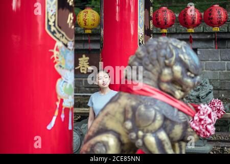 Donna asiatica sorridente positiva in camicia casual appoggiata sulla colonna Mentre si passa una giornata di sole nel tempio della religione buddista tradizionale contro Monumento del leone a Taipei Foto Stock