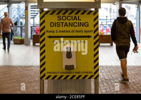 gel di mani igienizzante sotto un segno di allontanamento sociale al ingresso di un centro commerciale con persone che camminano oltre Foto Stock