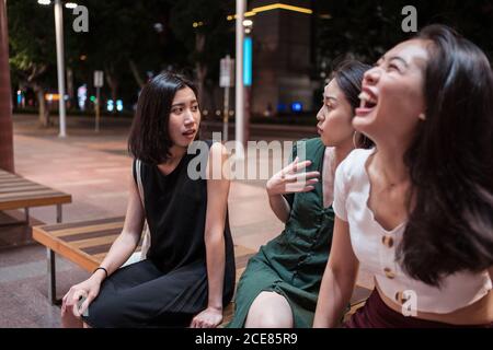 Compagnia di giovani girlfriends asiatici seduti su panca di legno e. parlare durante la passeggiata in città di notte mentre si guarda l'uno con l'altro Foto Stock