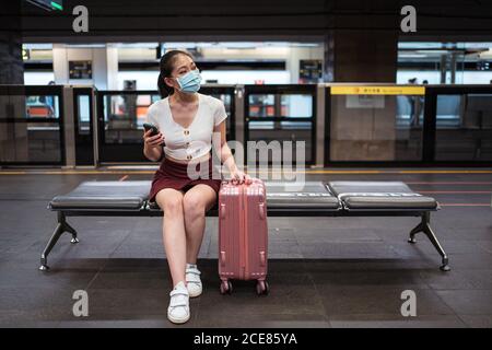 Giovane donna asiatica concentrata indossando mini gonna e maschera  facciale navigare sul telefono cellulare mentre si è seduti sulla panca con  la valigia in stazione ferroviaria Foto stock - Alamy