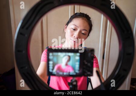 Messa a fuoco giovane Asian Donna stetting fotocamera su treppiede con anello luminoso mentre si preparano per la creazione di nuovo blog di bellezza a casa Foto Stock