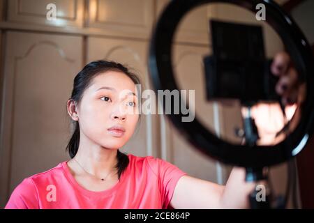 Messa a fuoco giovane Asian Donna stetting fotocamera su treppiede con anello luminoso mentre si preparano per la creazione di nuovo blog di bellezza a casa Foto Stock