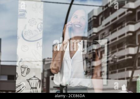 Donna elegante in abbigliamento formale che parla sul telefono cellulare mentre in piedi vicino a una parete di vetro nel caffè e guardando lontano sorrisi