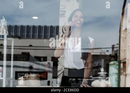 Donna elegante in abbigliamento formale che parla sul telefono cellulare mentre in piedi vicino a una parete di vetro nel caffè e guardando lontano sorrisi